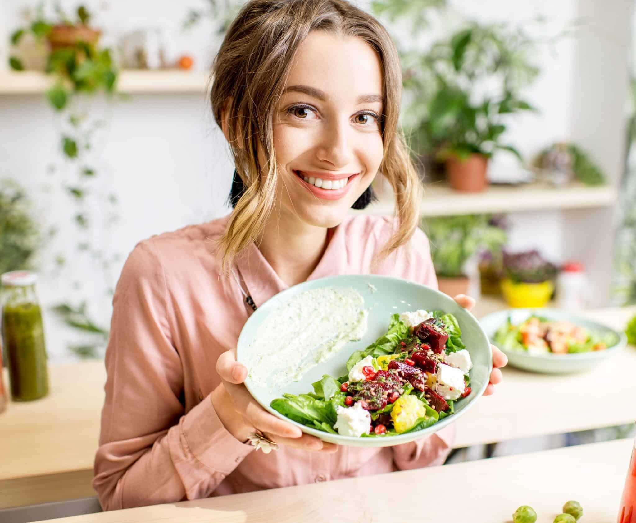 Portrait of a young woman holding a plate of salad sitting indoors surrounded with green flowers and healthy vegan food